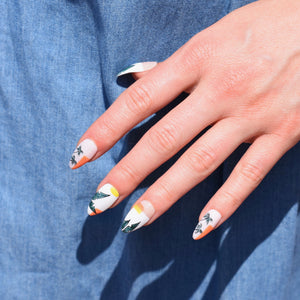 Hand-painted gel polish nail art press-ons in an almond shape.  Tropical nail art with some palm leaves and palm trees are scattered on the nails.  The base color of the nail design is a soft pink with modern color blocking accents in a golden yellow and warm orange.   All nails have a glossy finish.  One had is pictured over a denim backdrop.   