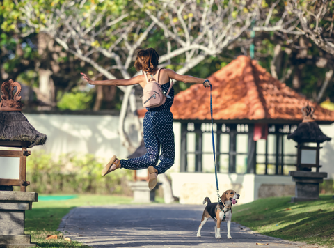 Woman leaping in the air facing away from the camera with her arms spread wide holding a leash with a beagle standing below