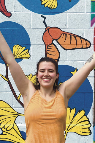 Photo of smiling young woman with light skin and brown shoulder-length hair. She is in front of a painted mural and has her arms outstretched upwards.