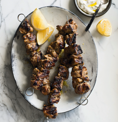 Close up of lamb skewers with lemon wedges and tzatiki on a light marble table