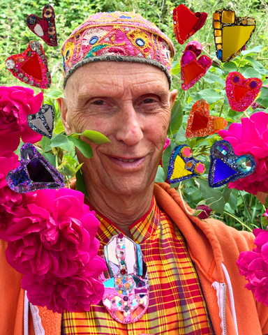 Sculptor Andrew Logan photographed in a beautiful rose garden, surrounded by hot magenta pink roses and a selection of his hand-made, colourful heart brooches.