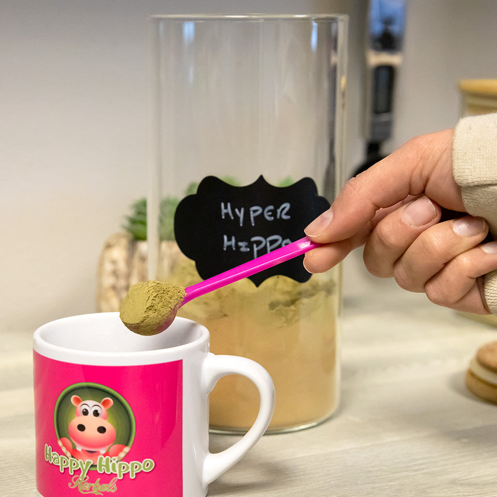 Photographic image depicting a happy hippo brand cup, being filled with Green Maeng Da Kratom powder to make kratom tea; A well manicured hand fills the cup uses a 1 gram little pink measuring scoop to fill the cup with kratom powder.