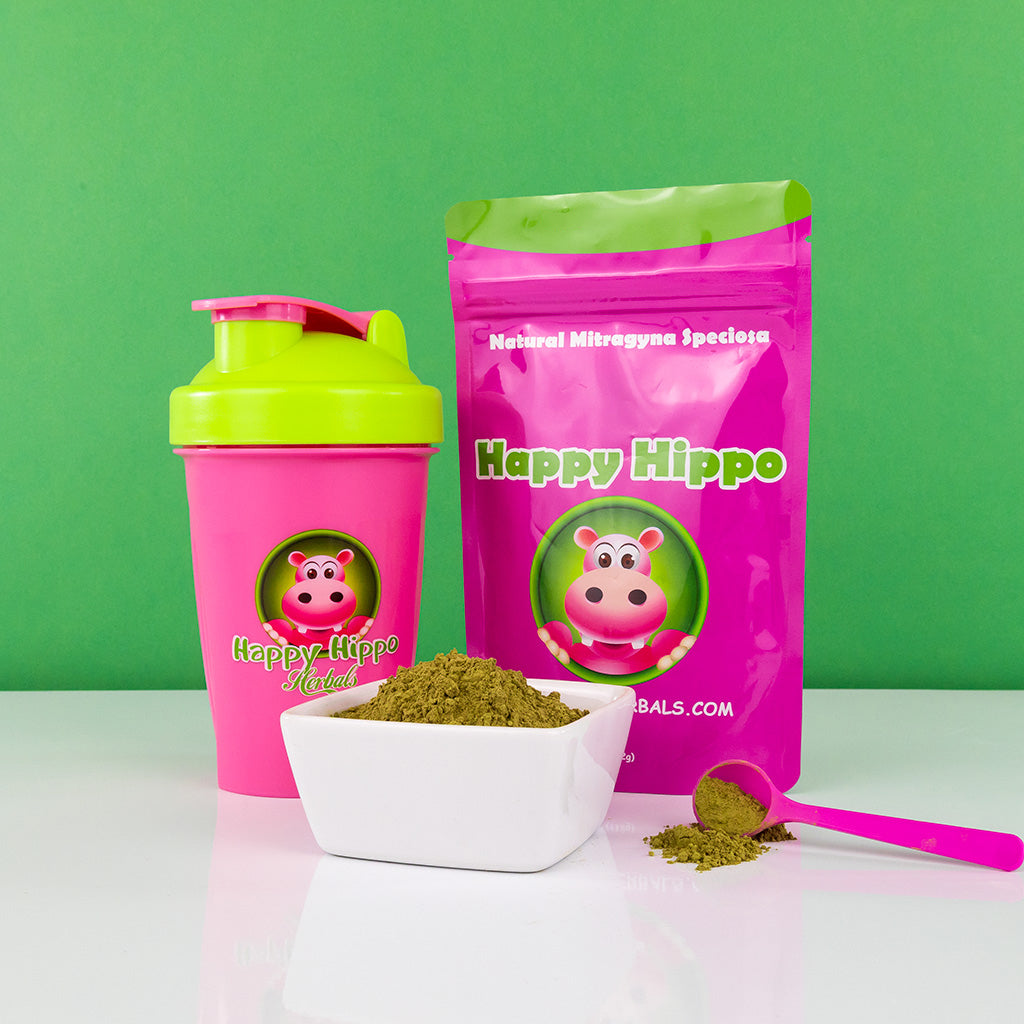 Photographic image depicting a 16oz shaker cup, as well as a branded packet of happy hippo Green Borneo kratom powder. A white bowl, as well as a 1oz little pink measuring scoop - both heaped with kratom powder, occupy the foreground.