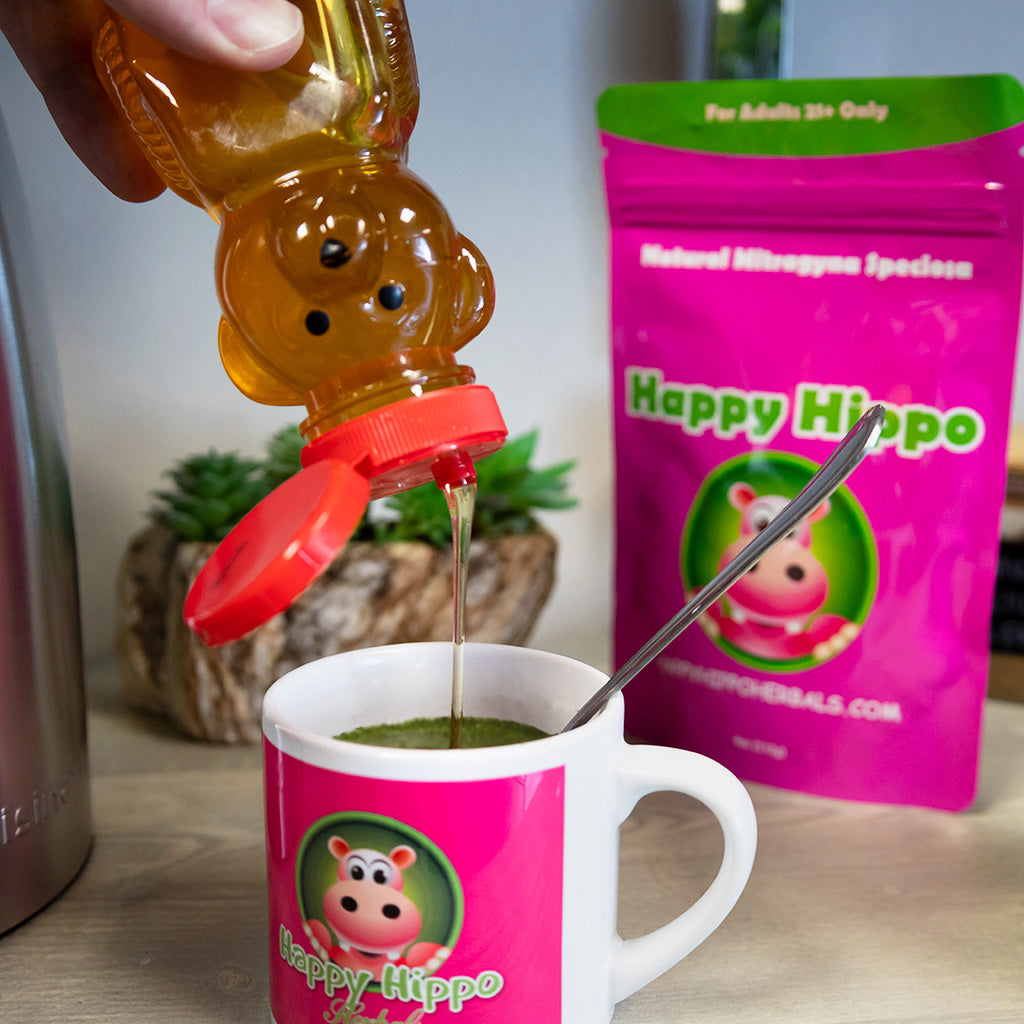 Photographic image depicting a Happy Hippo branded coffee cup filled with kratom tea. Above the cup, a pair of hands squeeze a plastic honey-bear bottle sending a stream of golden honey into the cup of kratom tea as a sweetener.