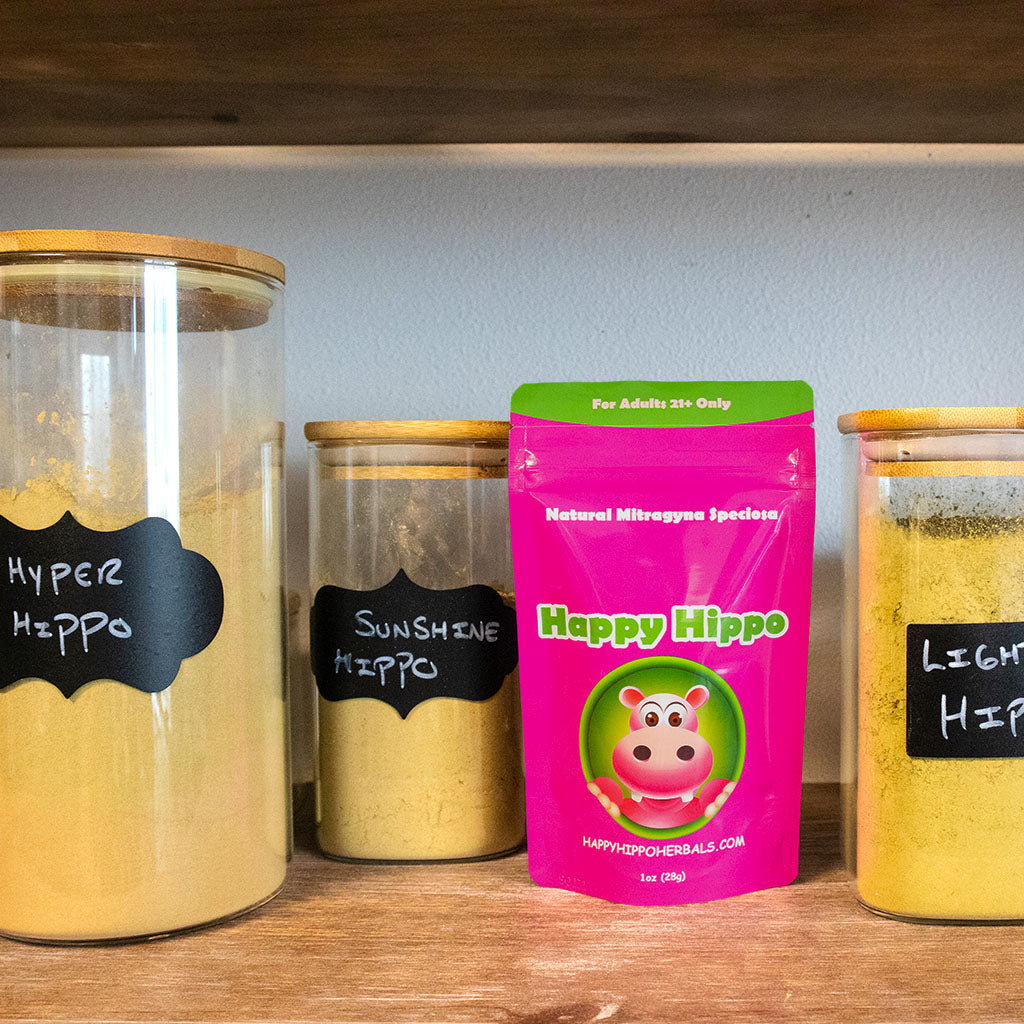 A photographic image depicting a shelf filled with glass jars containing different strains of kratom powder; The shelf includes Green Maeng Da (Hyper Hippo), Yellow Maeng Da (Sunshine Hippo), White Thai (Lightning Hippo), as well as a packet of Happy Hippo Brand Red Vein Bali (Red Dragon) Kratom Powder
