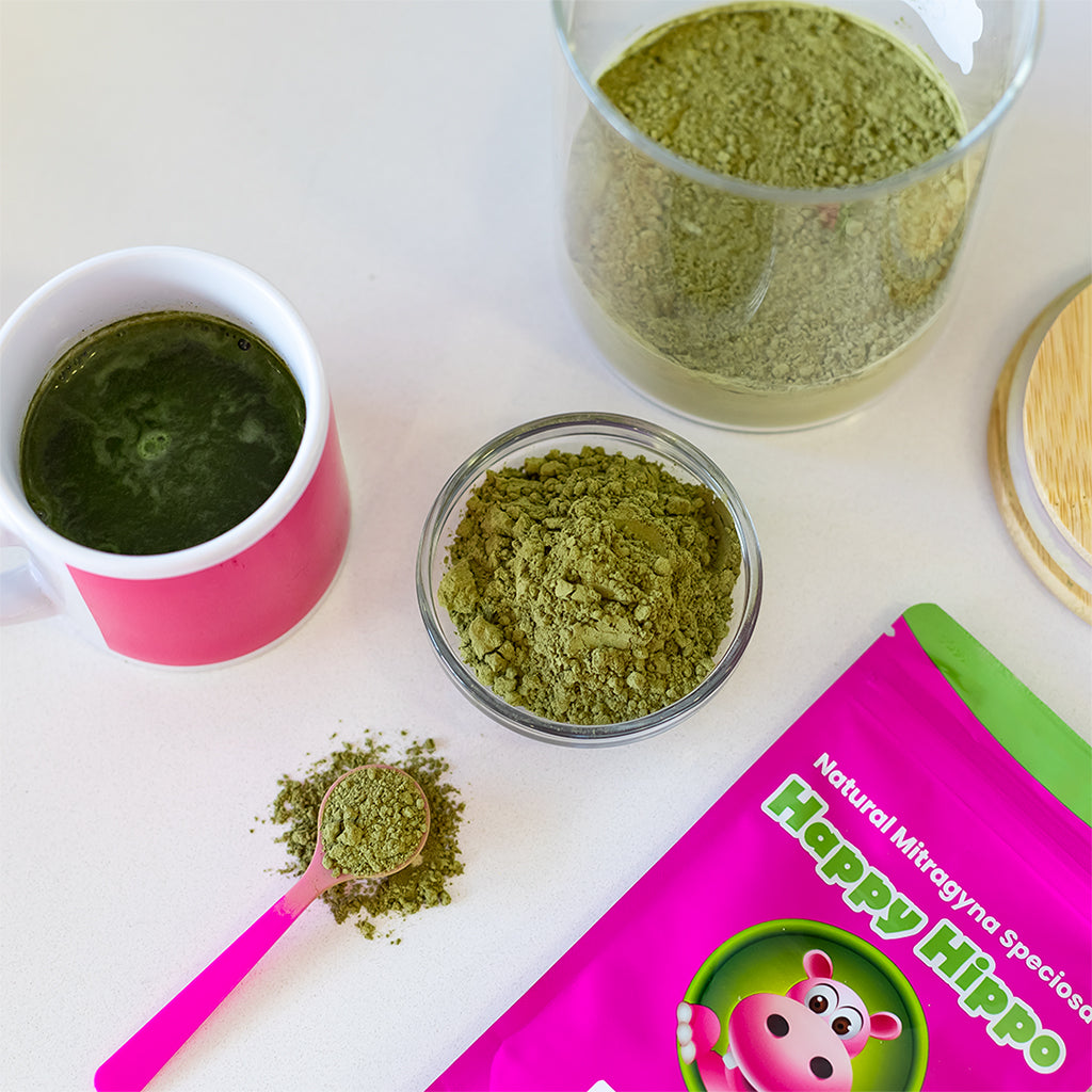Photographic image depicting a variety of Happy Hippo brand kratom powders, along with a warm cup of kratom tea, and a little pink measuring scoop, heaping with kratom powder.
