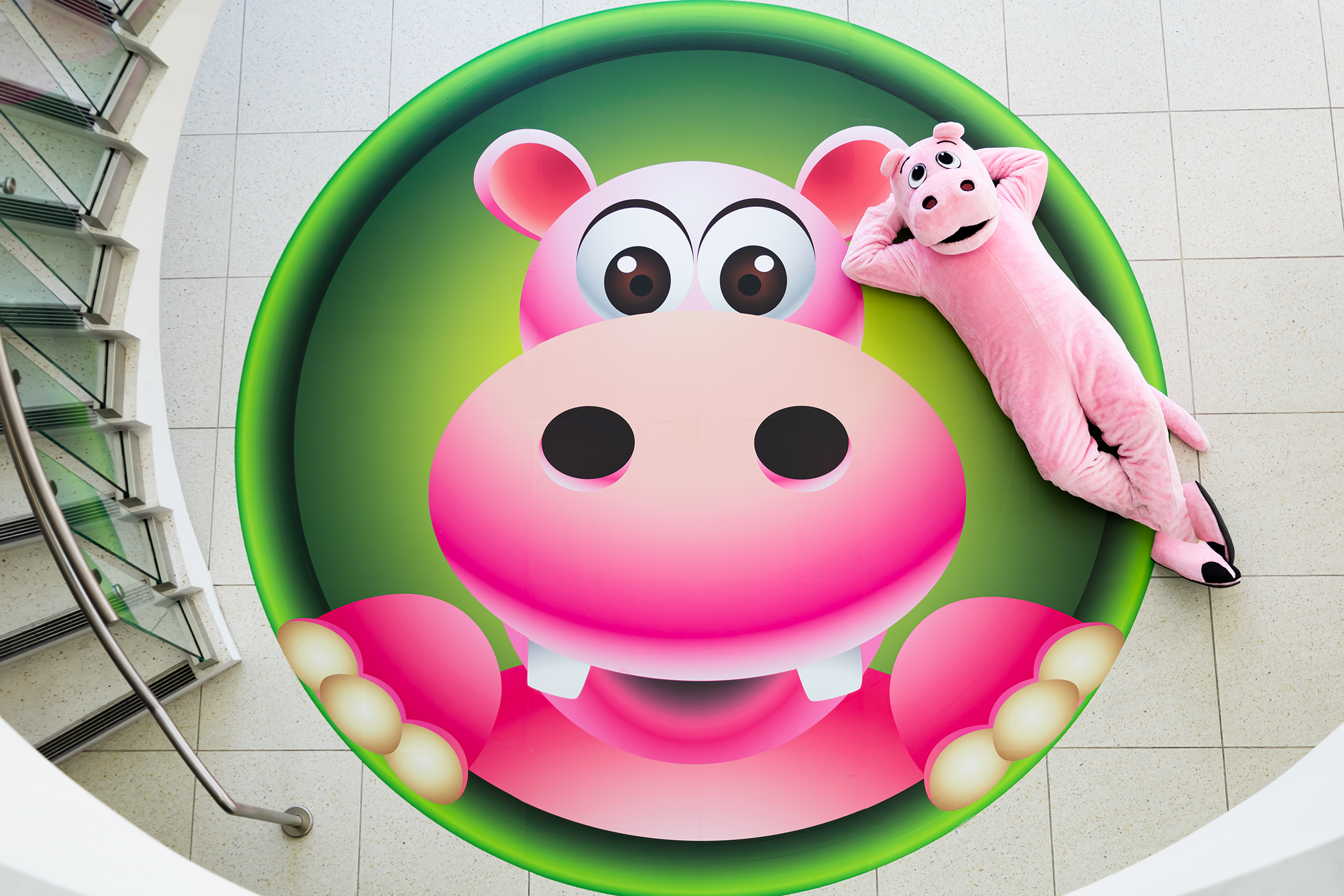 Large Happy Hippo logo on the ground with a person in a pink Hippo costume lounging next to it. 