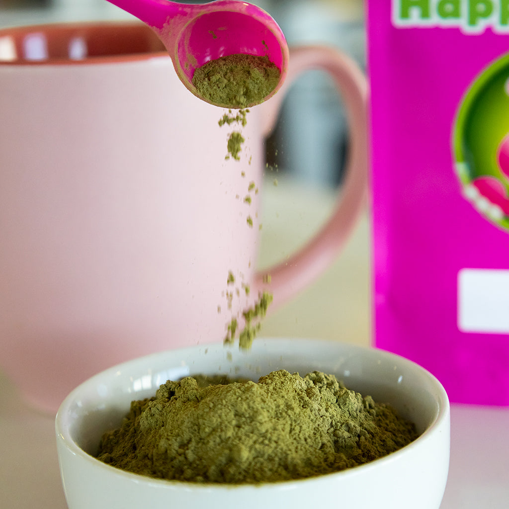 Photographic image depicting a small ramekin being filled with Happy Hippo branded loose white maeng da kratom powder, from a 1-gram, little pink measuring scoop.