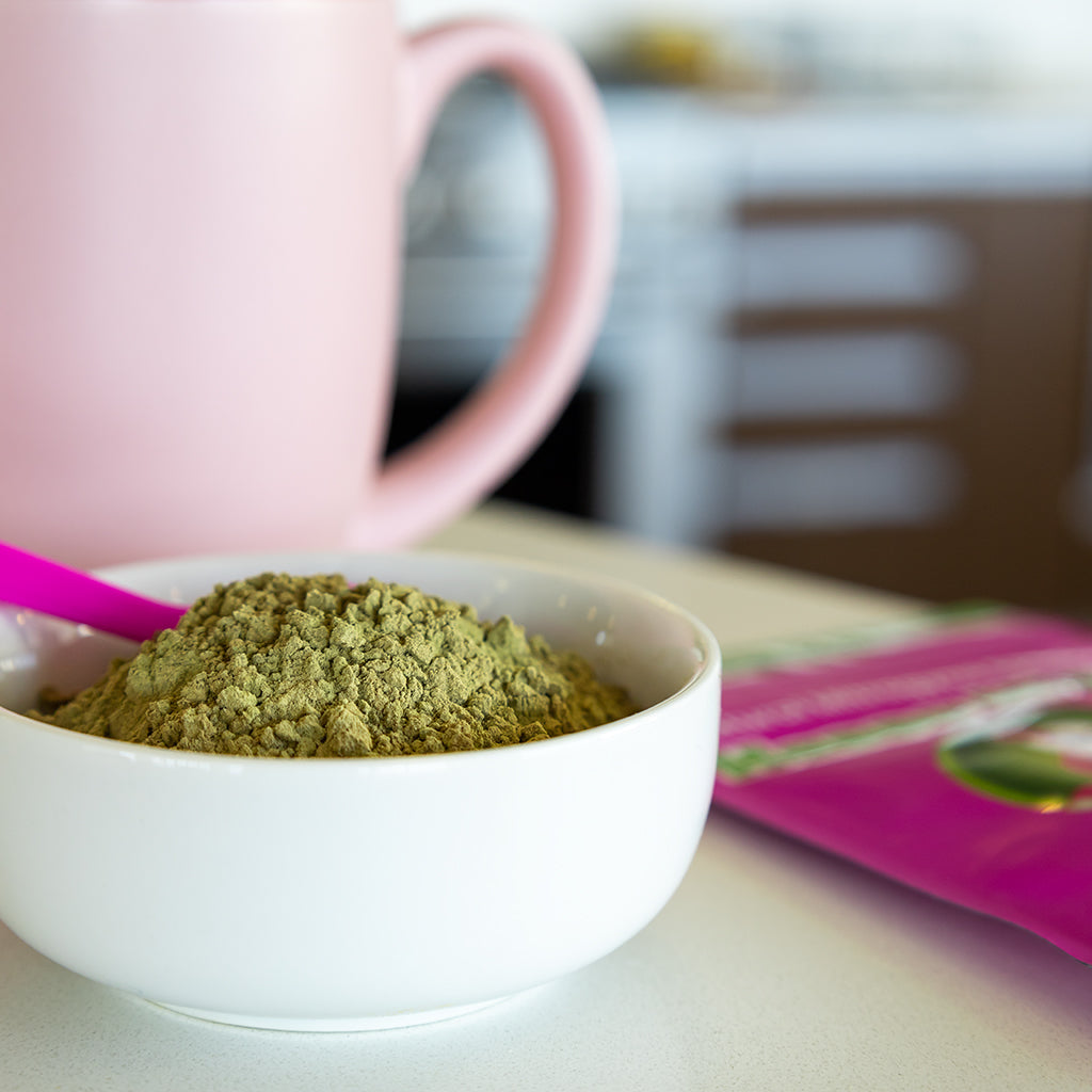 Photographic image depicting a bowl of loose Malay Kratom Powder sitting in the foreground, with a branded packet of Happy Hippo Malay Kratom and a pink kratom tea cup, blurred in the background.