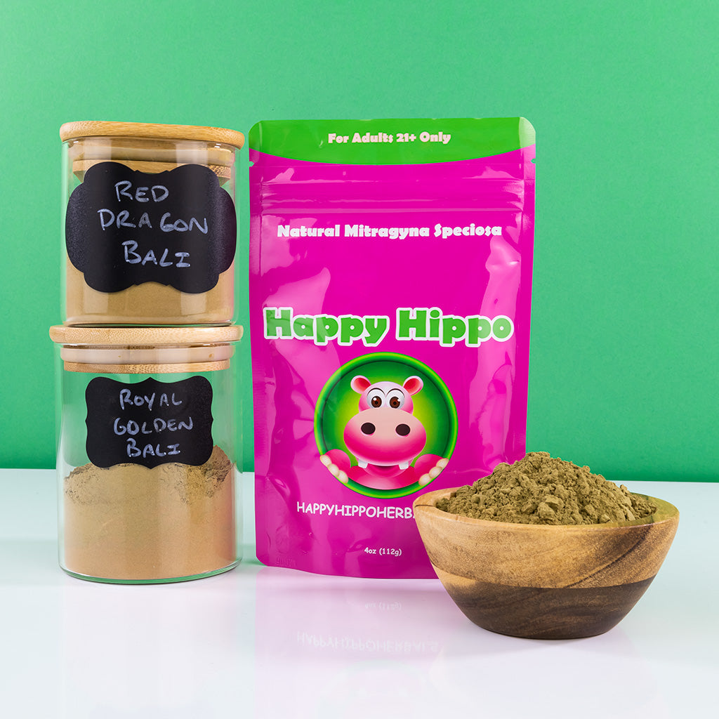 Photographic image depicting a 4oz packet of Happy Hippo Brand kratom powder, sitting next to a wooden bowl filled with a heaping mound of loose Bali kratom powder; Sitting next to the kratom powder packet, are stacked two glass jars filled with kratom powder. The jars read: "Red Dragon Bali", and "Royal Golden Bali"
