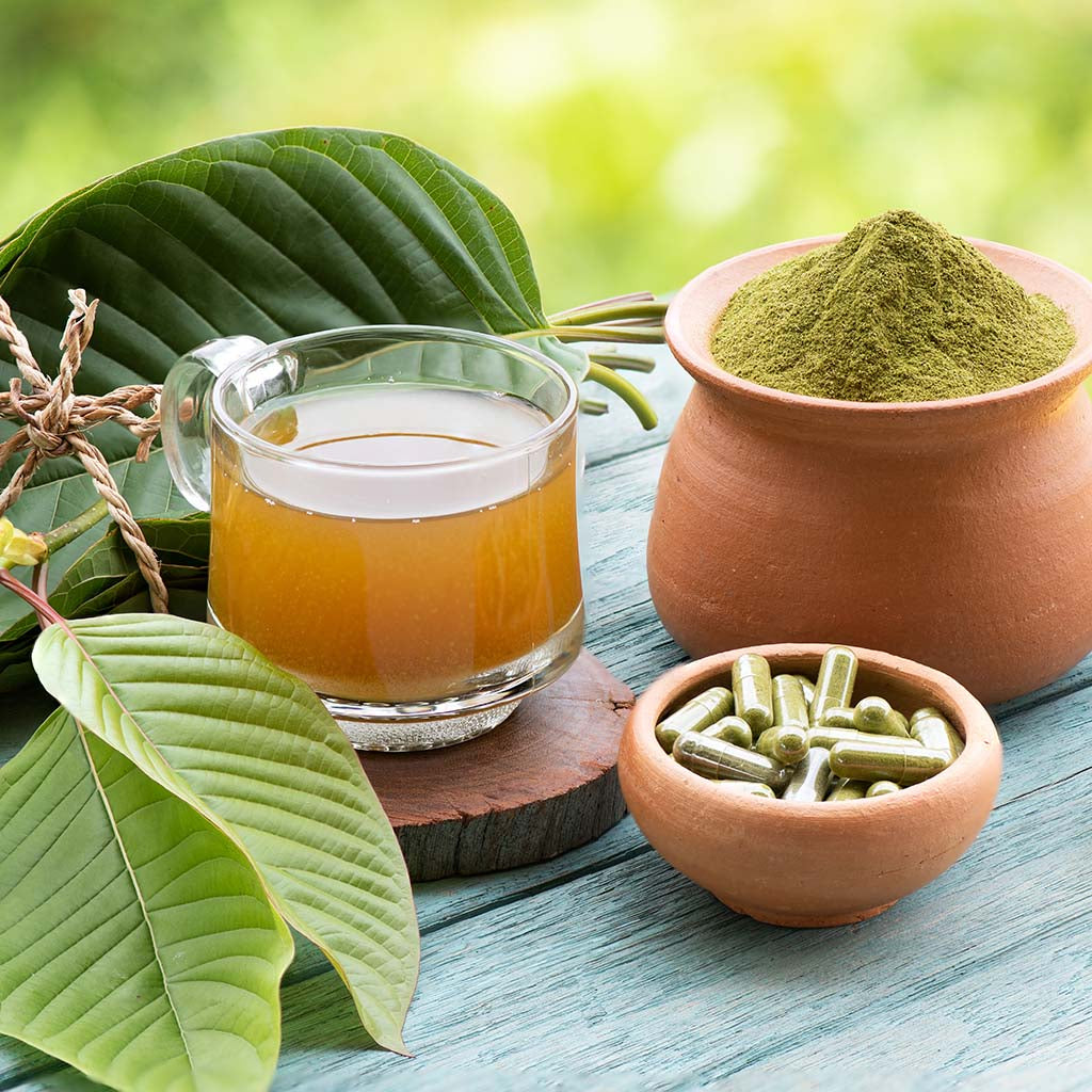 A photographic image depicting an array of botanical herbs, at the center of which are several kratom leaves, a cup of kratom tea, kratom powder, and a small dish containing a handful of kratom capsules