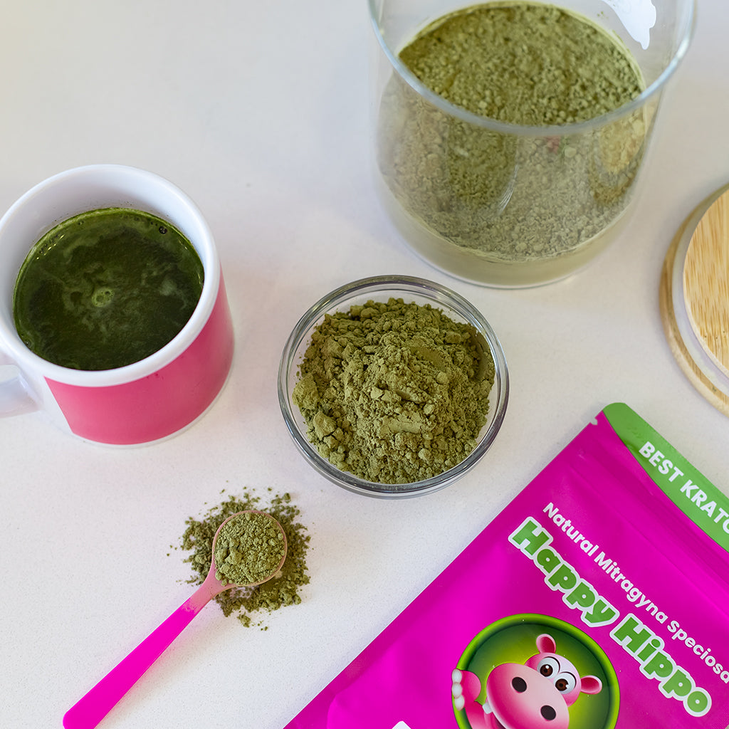 Photographic image depicting a top-down view of a coffee mug filled to the brim with warm kratom tea. Sitting next to the mug, is a glass ramekin filled with Green Maeng Da kratom powder, and a 1 Gram little pink measuring scoop.