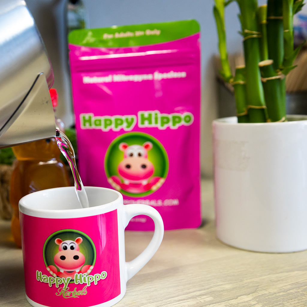 Photographic image of a happy hippo branded coffee cup being filled with hot water, in preparation to make kratom tea. In the back ground is a packet of Happy Hippo brand Green Maeng Da Kratom Powder, and a small, potted, rattan plant.