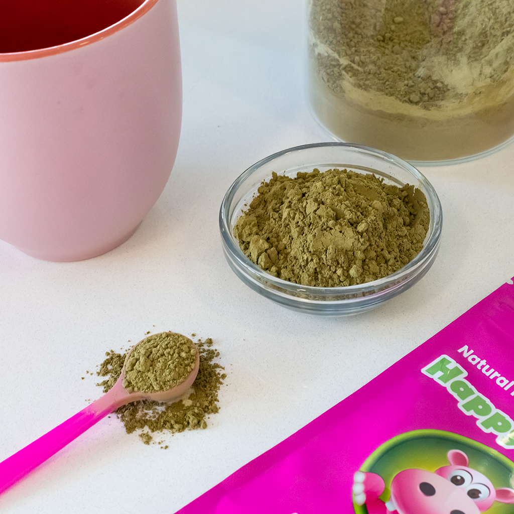 Photographic image depicting a teacup, and a few accessories (a 1-Gram pink measuring scoop, and a ramekin filled with loose red borneo kratom powder), next to which sits a Happy Hippo branded packet of Red Vein Borneo Kratom Powder.