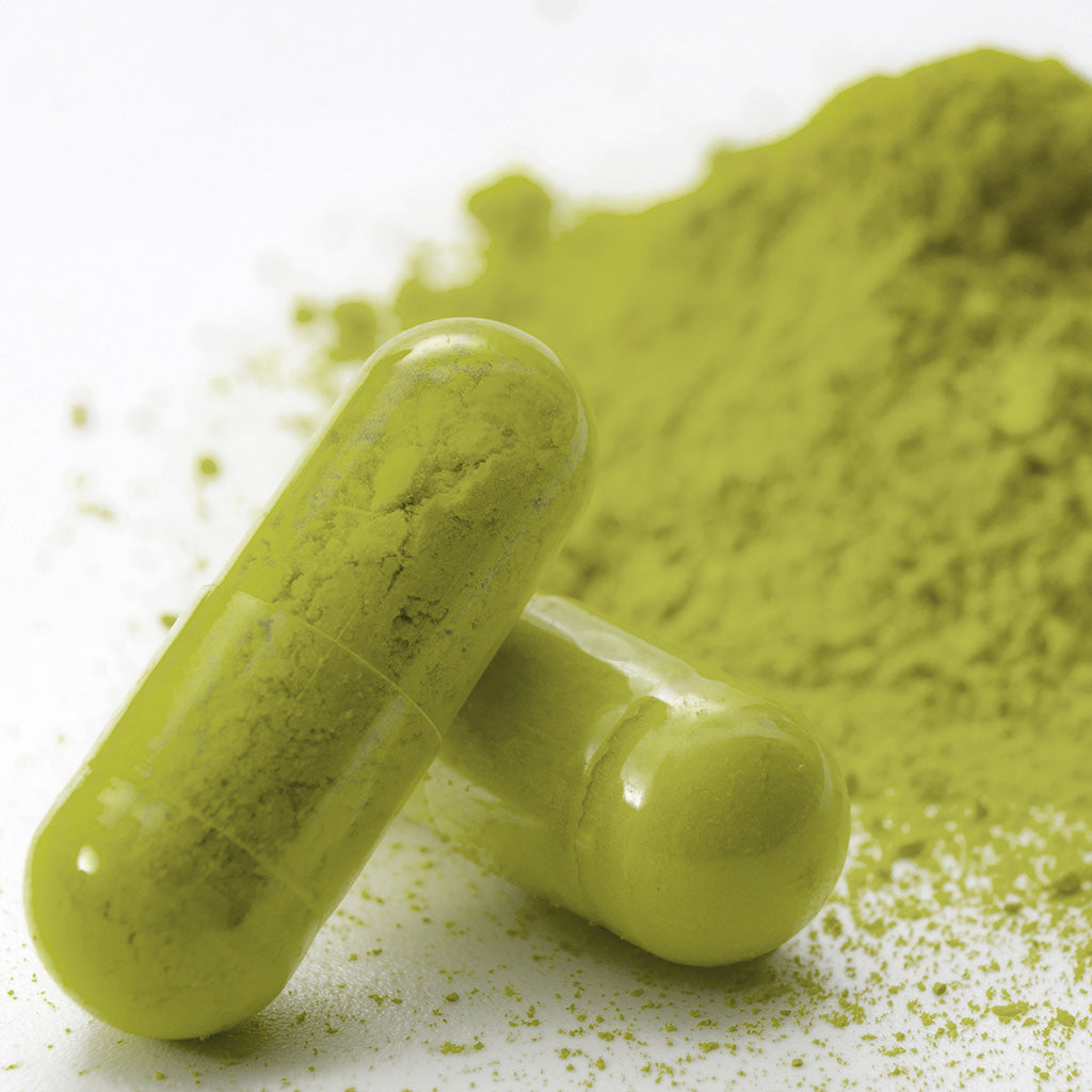 Photographic macro image depicting a close-up, detailed image of two white vein kratom capsules propped up in front of a stack of white vein kratom powder