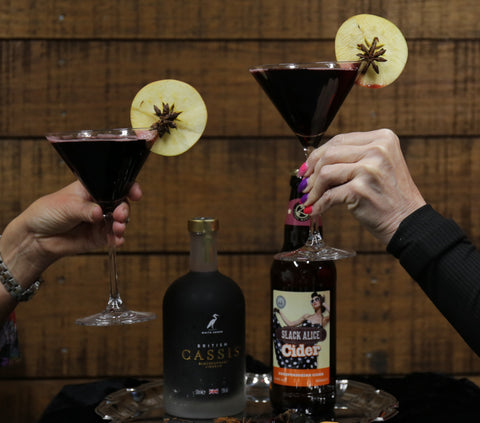 Image shows two hands holding martini glasses with theBlack Witch cocktail in, apple slices on each glass and a bottle of British Cassis and Slack Alice cider on a silver tray.