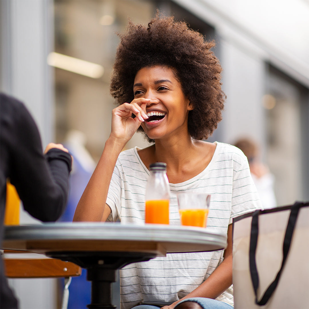 Woman of colour sitting at an outside table with another person. There is a juice bottle and glass of juice on the table and the woman is laughing / smiling / feeling good.