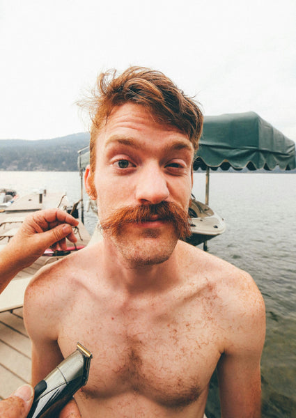 Shirtless man with uneven facial hair mid-trim making a funny face on dock by the water