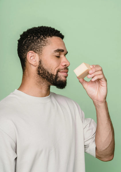 Bearded man in a light shirt smelling a bar of soap with eyes closed against a green background