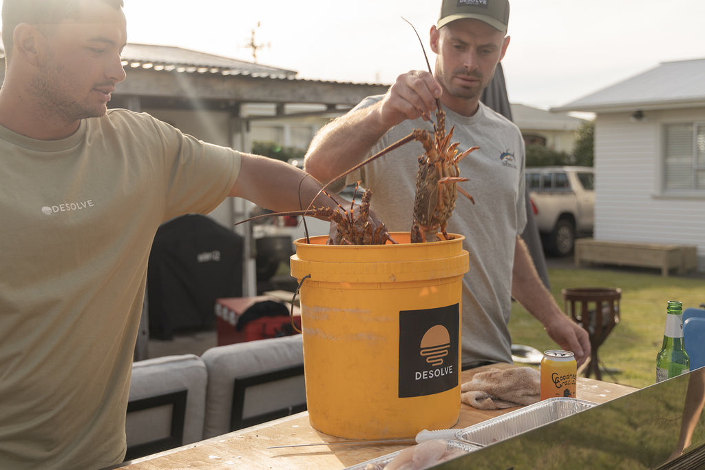 Puka and Bung getting the crayfish ready for the grill with his Goodness Gracious gin & juice to stay hydrated - wearing the Desolve Untouched Tee - Seagrass and the Albacares Tee - Grey Marle