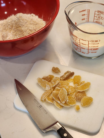 A bowl with the Gingerbread scone mix in it with crystalized ginger on a cutting board with a knife and a cup of whipping cream