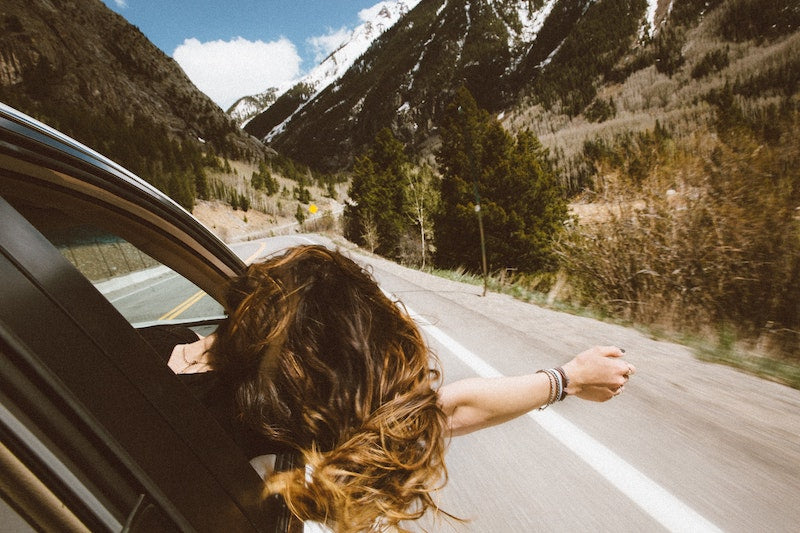 young woman in the passenger seat of a car with her head and hand out of the window