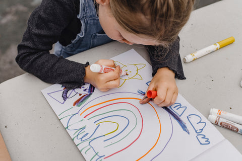 A small child using crayons to draw a colorful rainbow with a cat.