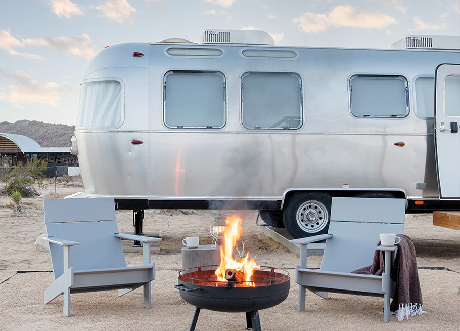 Image: Two Driftwood Lollygagger Lounge Chairs sit side by side behind a lit fireplace. The Lollygagger Lounge Chair on the right has a cup of coffee sitting on the right arm along with a folded blanket. Behind the chairs is an Airstream Trailer. This scene takes place in the desert at Autocamp in Moab, Utah