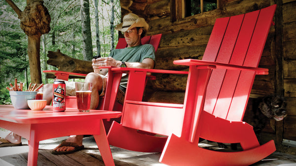 Image: A sideview of a man sitting on a cabin porch in an Adirondack Rocker Chair with another empty chair beside it.