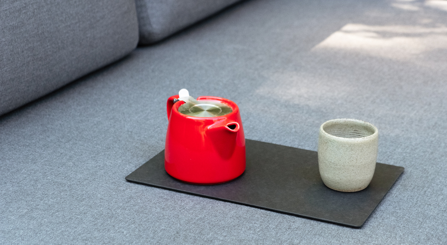 A red pot and a white cup of tea on a black cutting board, sitting on top of grey cushion