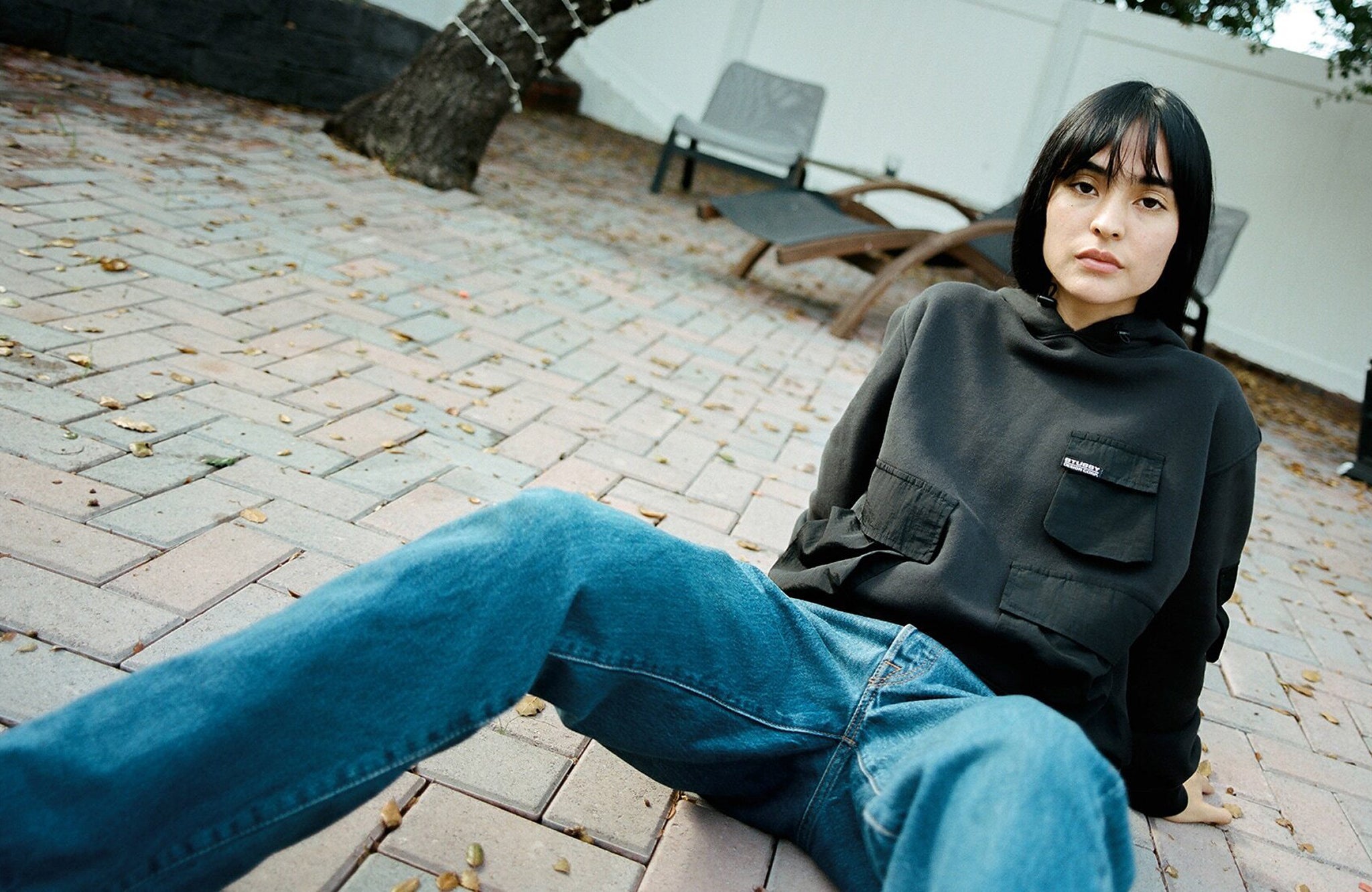 Female model sitting in on backyard stone pavement wearning military pocket hoodie and denim