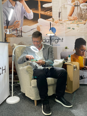 Brett sitting in an arm chair, using a magnifier and light to aid craft work.