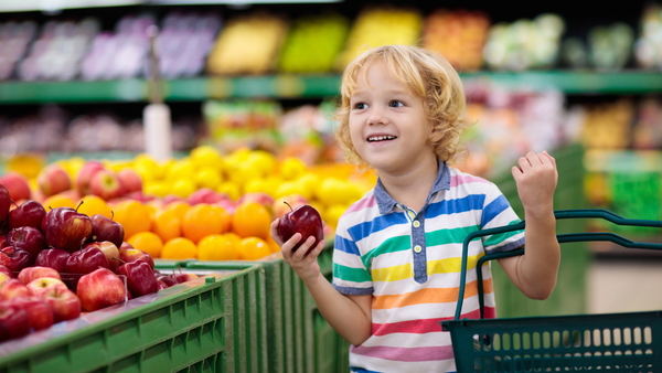 Junge steht im Supermarkt im gang. In der einen Hand hält er einen Apfel, in der anderen einen Einkaufskorb. Er ist stolz, dass er mit einkaufen gehen kann.