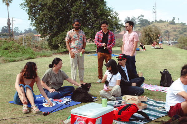A few friends at the Outdoor Asian x Rooted Fare picnic; some sitting on colorful picnic blankets and 3 people standing up, chatting.