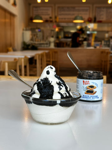Snow-white soft serve topped with Black Sesame Crunchy Butter on a white counter with Yang's Kitchen as background.