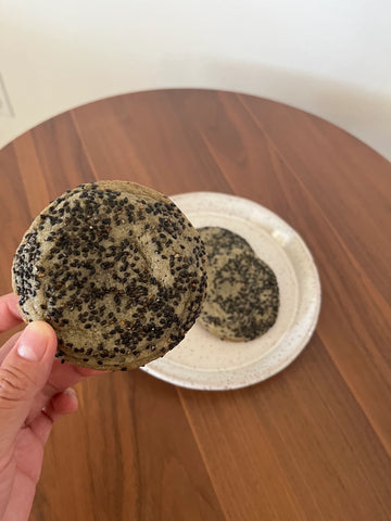 A black sesame cookie with black sesame seeds sprinkled all over is held up over a wooden table. Photo courtesy of Batter Together.