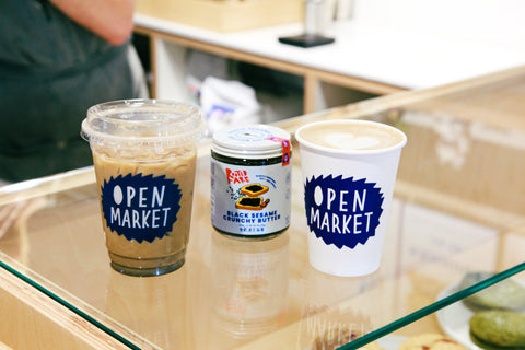 Two Open Market Black Sesame Lattes on a glass counter with a jar of Black Sesame Crunchy Butter in the middle.