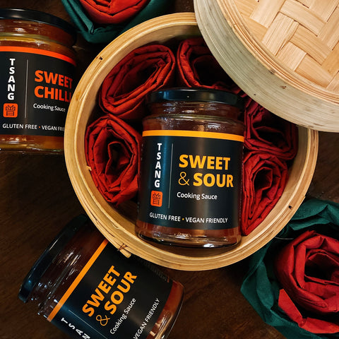 Jars of Tsang Sauce in Sweet Chilli and Sweet and Sour flavour placed inside and around a bamboo basket set on a wooden table