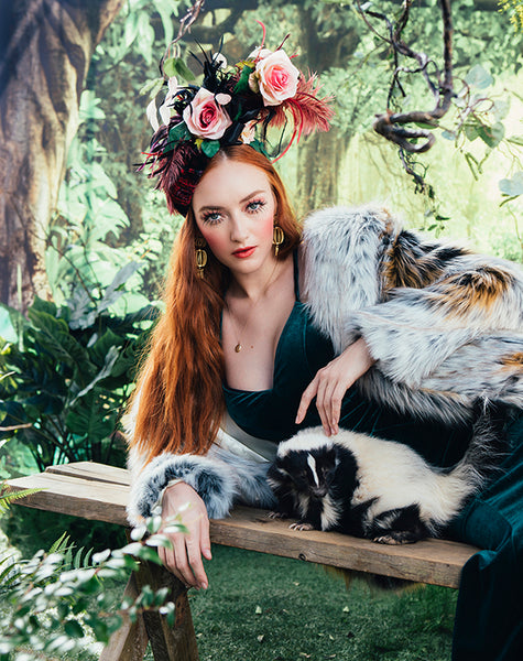 woman in teal gown lying on bench wearing faux fur coat petting a skunk