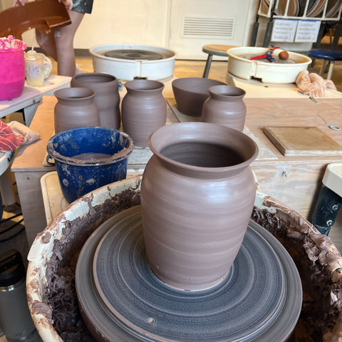 Photo of a vase on a pottery wheel. It is surrounded by several other finished vases. All of them are dark brown