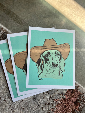 Photo of three prints by the artist of a dachshund in a cowboy hat on