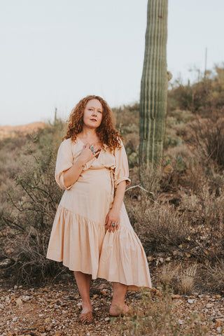 Photo of the artist, a red-haired woman in a peach dress in front of a desert background with a saguaro cactus