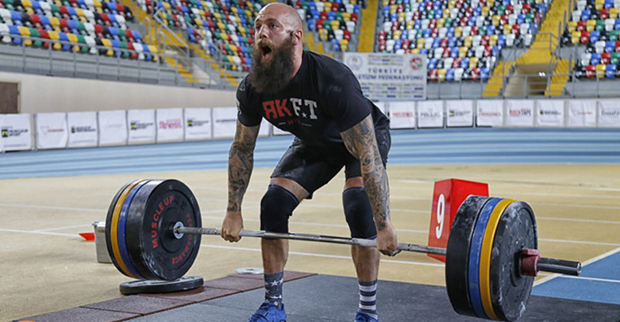 Bald, bearded man with tattoos exerting effort while deadlifting heavy weights in a weightlifting competition, with empty stadium seats in the background.