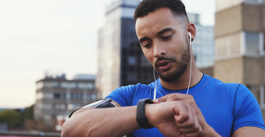 Focused man in a blue shirt checking his fitness tracker while wearing earphones, with urban buildings in the soft-focus background.