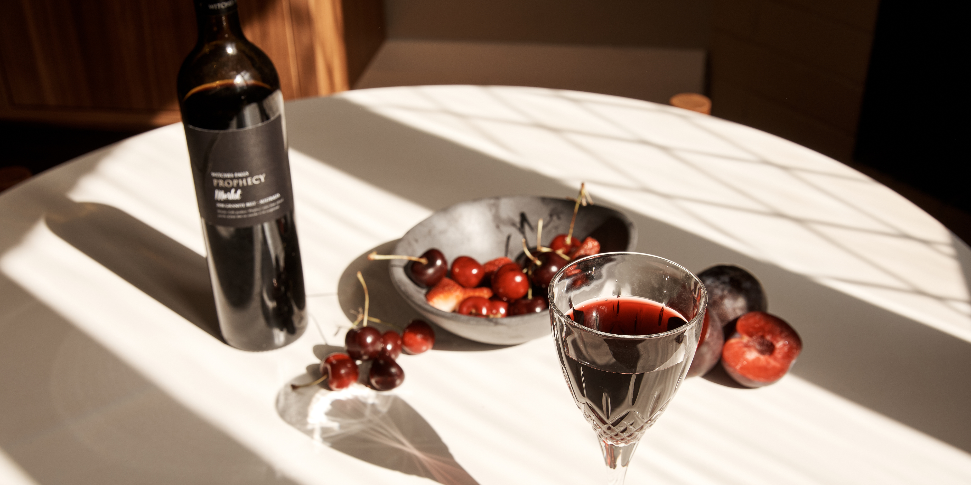 A bottle of Witches Falls Prophecy Merlot on a round table with a glass of red wine, and a bowl of red cherries and plums