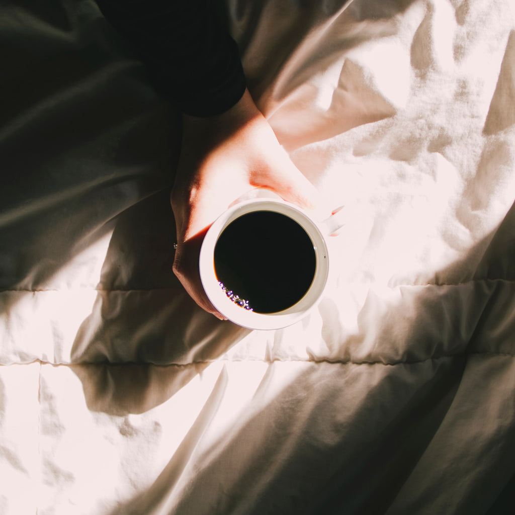 Kahvikuppia pitelevä käsi auringon valaisemalla sängyllä.