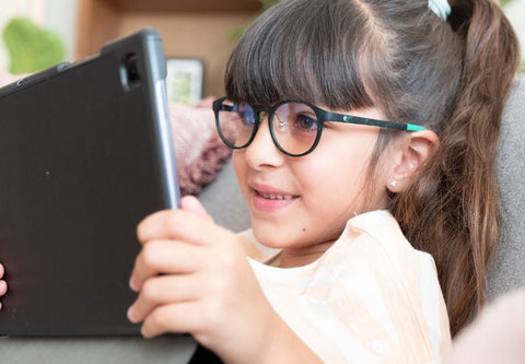 Ein kleines Mädchen schaut mit Brille auf ihr Tablet auf dem Sofa