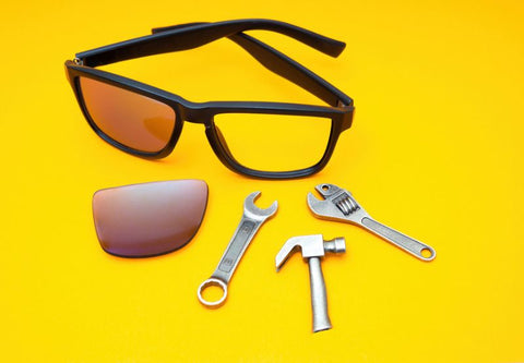 Sonnenbrille mit einem fehlenden Glas und einem Glas daneben auf gelbem Hintergrund mit Ersatzwerkzeugen
