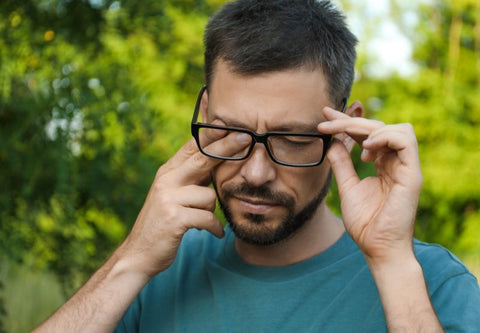 homme avec des lunettes qui se gratte l'oeil sur fond de verdure