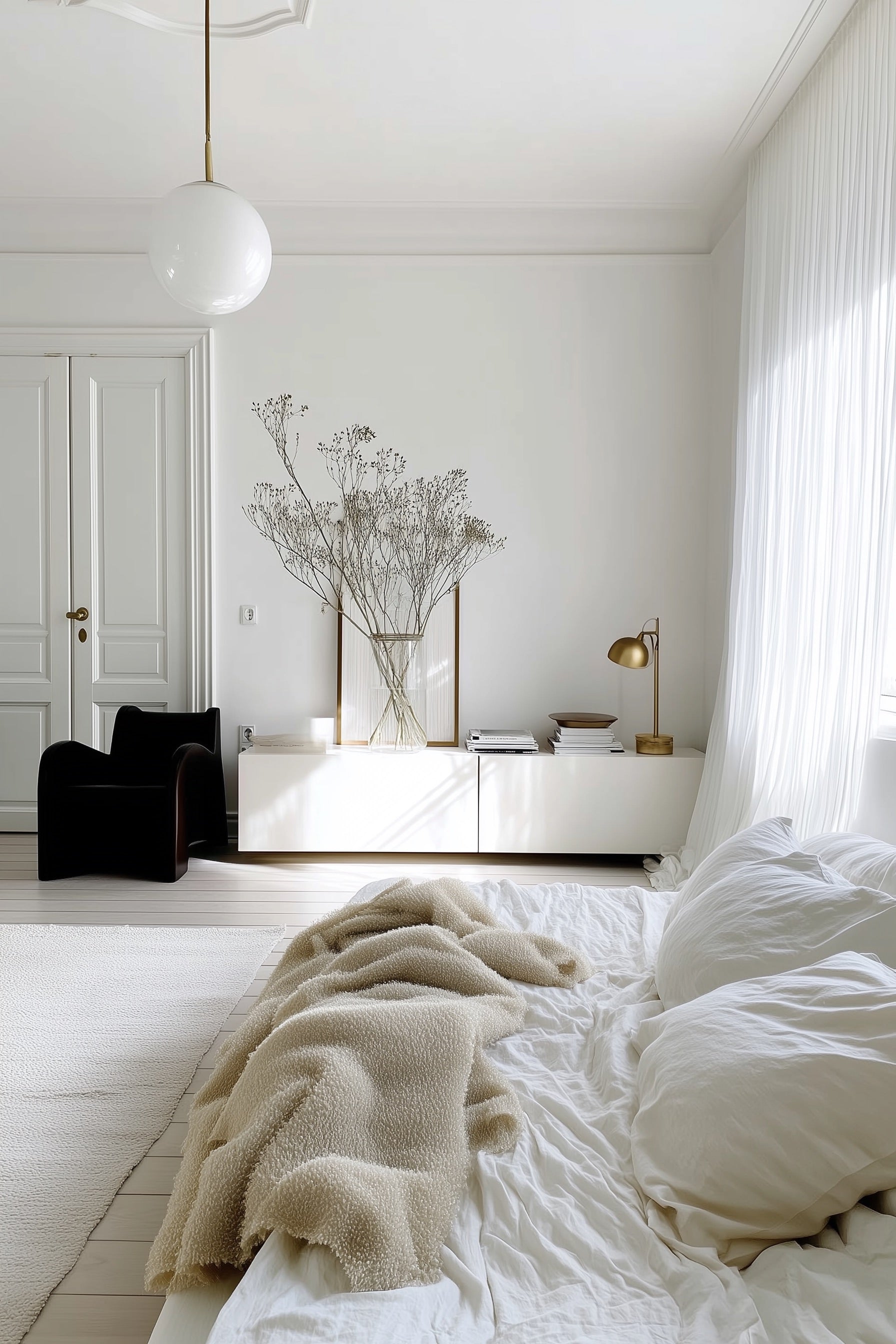 A serene bedroom with white walls, soft bedding, a beige textured throw, a modern black chair, and a low cabinet styled with books, a brass lamp, and a large vase of dried branches